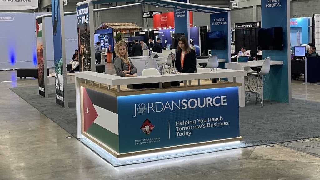 A trade show booth labeled Jordan Source features two women standing behind a counter with the Jordanian flag and the slogan Helping You Reach Tomorrow’s Business, Today! Several other booths are visible in the background.