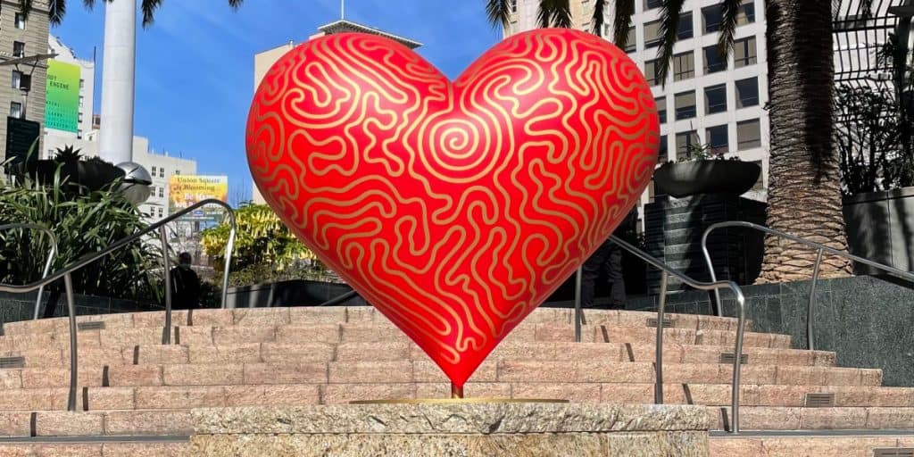 A large red heart sculpture with swirling yellow patterns stands on a stone pedestal in an outdoor plaza in downtown San Francisco, surrounded by plants, stairs, and tall buildings.