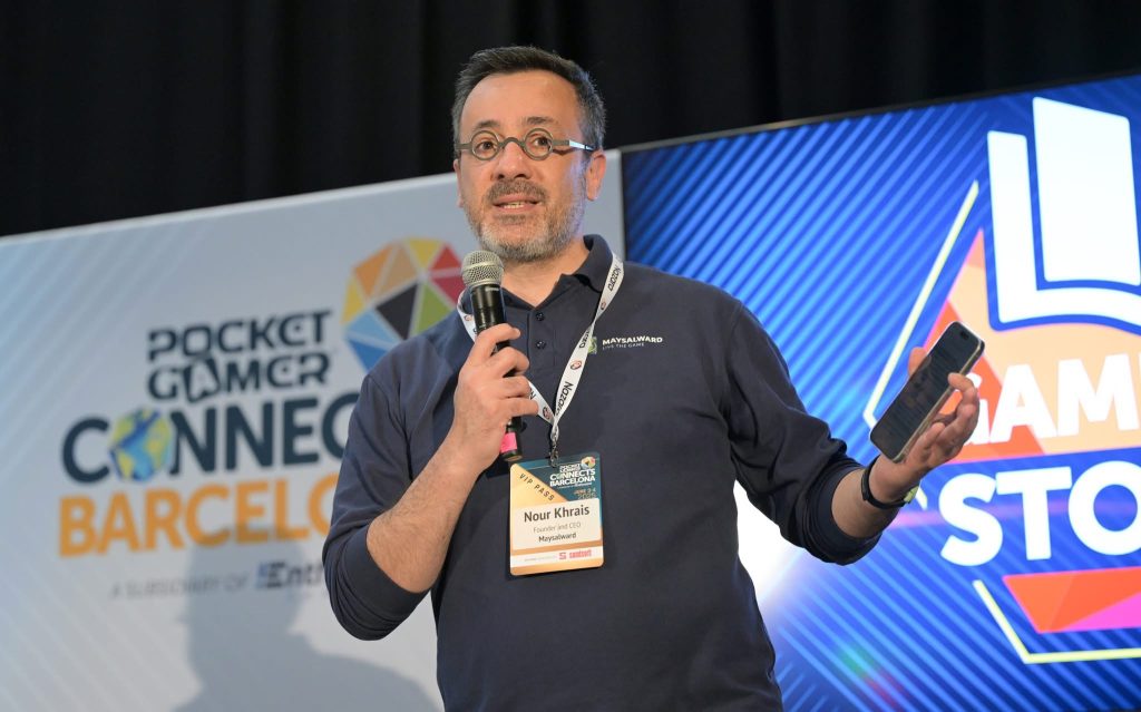 Nour Khrais with glasses holds a microphone and a smartphone, speaking on stage at Pocket Gamer Connects Barcelona. He wears a lanyard and a navy shirt, with conference branding displayed on screens behind him.