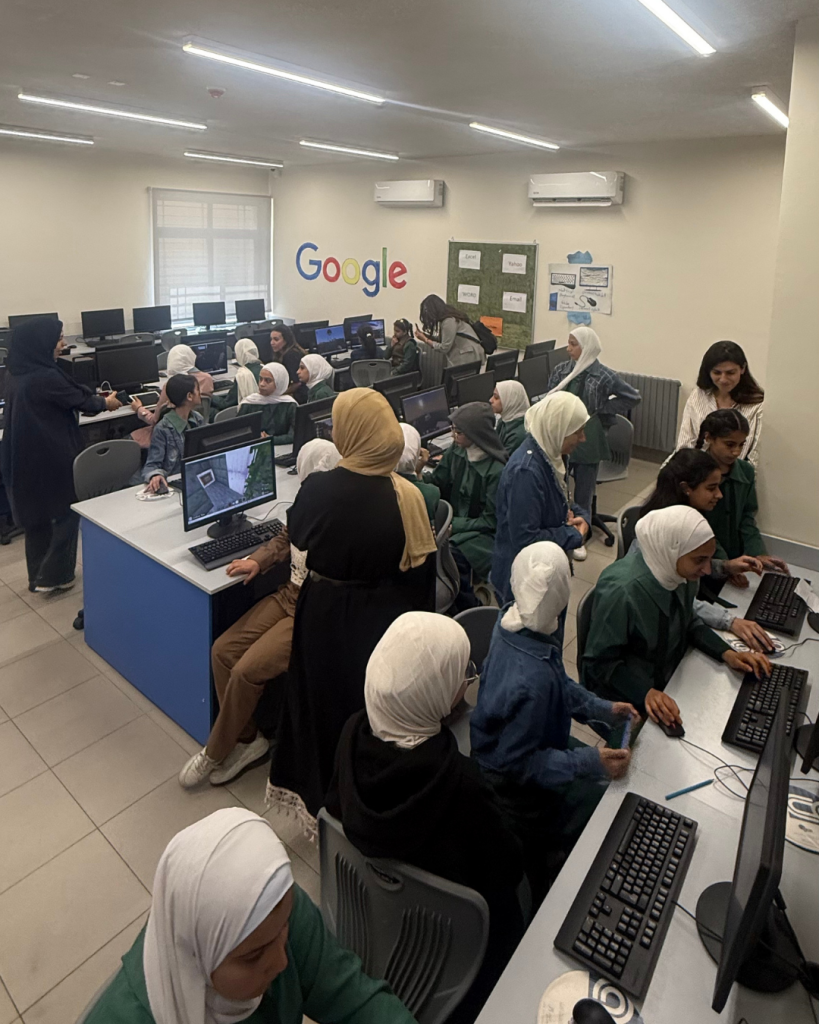 A classroom filled with students, mostly girls wearing hijabs, working on desktop computers. Several adults supervise. The word Google is displayed on the wall in colorful letters.